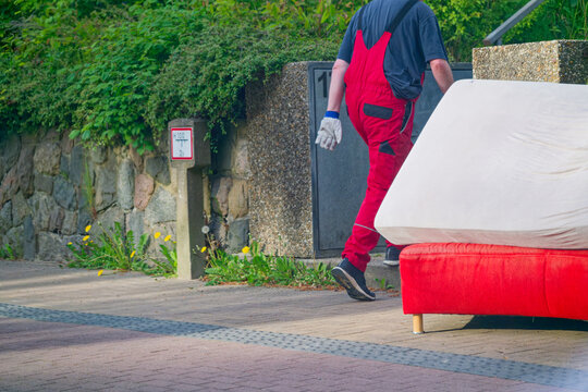 Worker in red overalls carrying a mattress on a sidewalk near a stone wall and greenery