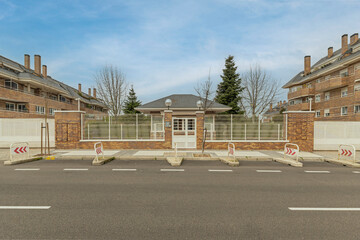 Asphalt road with parking bollards in front of a brick wall and white fence with a central gatehouse, flanked by large brick apartment buildings. © Toyakisfoto.photos
