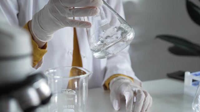 Unknown scientist wearing white protective gloves is carefully shaking up a clear chemical solution inside an Erlenmeyer flask in a laboratory, close up. Science and medicine