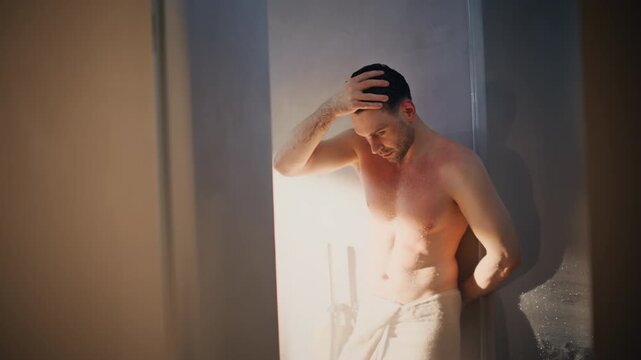 Shirtless man leaning shower wall in white towel. Wet male model relaxing spa