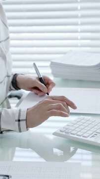 Financial professional performing calculations and writing data, preparing tax forms, managing budget, or completing administrative tasks on a bright office desk with stacks of documents