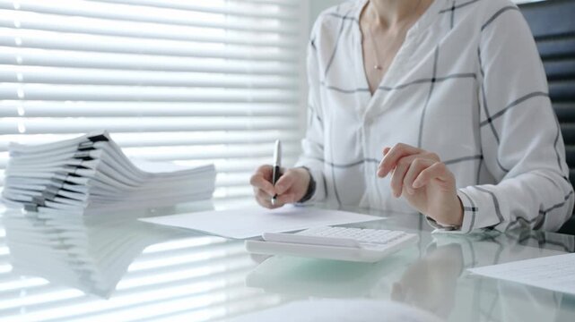Woman's hands using a calculator and holding a pen, working with financial documents and a pile of papers on a clear desk in an office setup