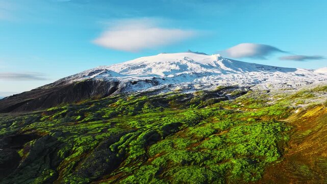 Moss covered volcanic plateau rising toward snow capped stratovolcano beneath clear blue sky with lenticular clouds and rugged lava cliffs in remote alpine wilderness