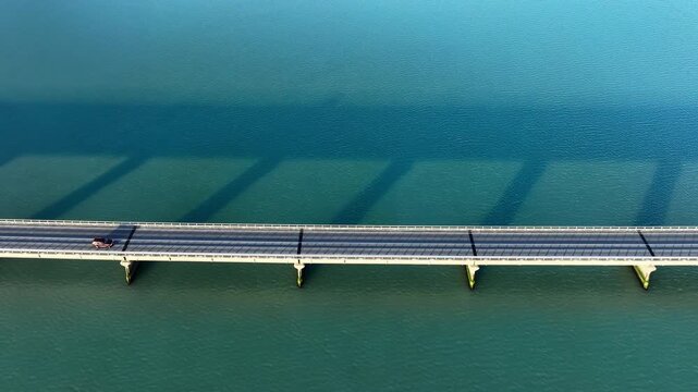 Aerial bridge crossing calm teal water with long diagonal pier shadows and clean geometric lines stretching across wide open landscape, with bridge, aerial, topdown, pier detail