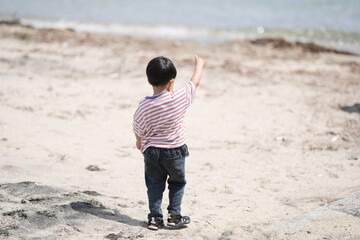 Child stands on sandy beach, pointing toward water. Back view shows curious, focused posture. Sunlight glows softly over calm shoreline. Perfect for family, vacation, or childhood adventure themes © Masakazu Tokashiki