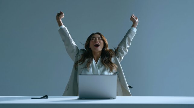 Businesswoman celebrates success with energetic arm raise, expressive joyful smile, wearing suit beside laptop on modern desk against vibrant solid background.