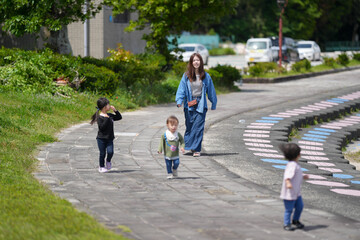 Fototapeta premium Young children play on a curved stone path. Woman walks nearby, smiling gently. Green grass and trees frame the scene. Bright sunlight enhances the peaceful mood. Perfect for family, education