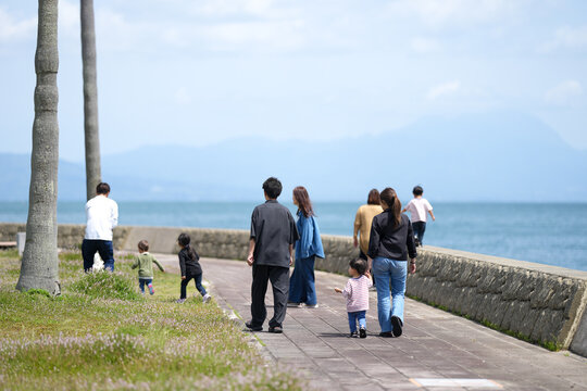 Family strolls along seaside path. Sky is soft, sea stretches far. People walk side by side, relaxed. Children laugh, exploring grass. Mountain range frames distant horizon. Ideal for family, travel