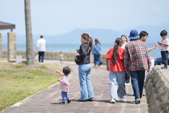 Sunlit seaside path, children and adults stroll together. Little girl points toward ocean, eyes filled with wonder. Group chats, laughs, and casual steps follow along. Coastal breeze whispers