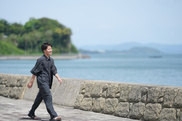 Man walks calmly along seaside stone path. He smiles, enjoying quiet coastal air. Background shows gentle waves and distant green hills. Ideal for travel, wellness, or calm lifestyle themes © Masakazu Tokashiki