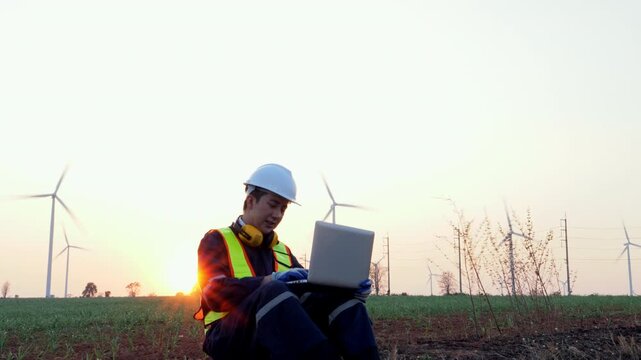 Professional man intently works on laptop outdoors during stunning sunset beside modern wind turbines, evoking industrious energy and utilizing technology's potential during golden hour light.