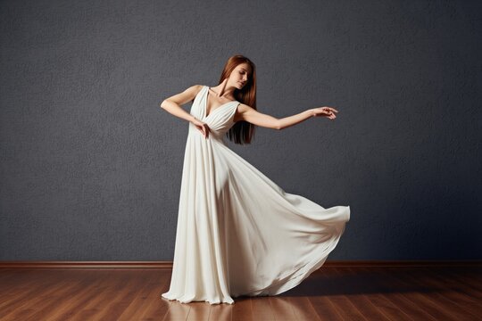 Young woman with long red hair gracefully dances in flowing white gown. Moves elegantly in studio setting against textured grey wall, arms extended in fluid motion. Floor polished wood, reflecting
