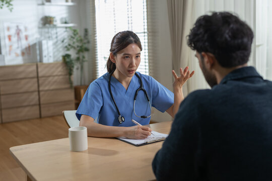 Asian female nurse talking to male patient during serious health consultation in bright clinic room holding pen and clipboard wearing blue uniform and stethoscope providing expert medical care