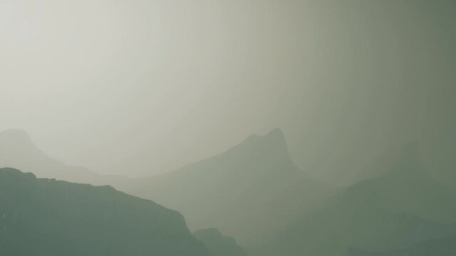 Early morning fog envelops mountains, making them appear soft and undefined. The light is dim, creating a sense of mystery in the landscape. Nature displays its power and beauty.