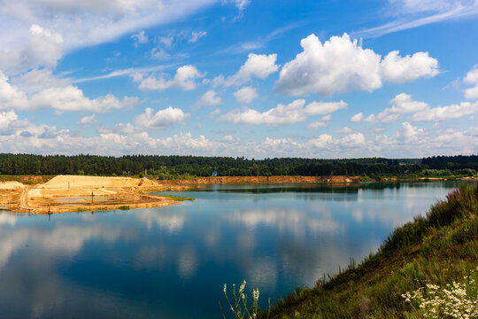 Serene turquoise water fills an abandoned sand quarry reflecting fluffy white clouds under a bright summer sky with a distant forest rimming the picturesque industrial landscape