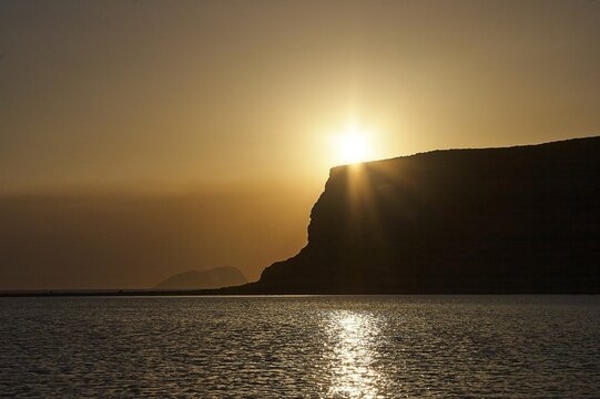 Dusk, sunset, sun as star, sun behind hill, bronze sky, cliff, island, sun reflection on water, Gramvoussa peninsula, Pirate's Bay, Balos, Tigani, West Crete, island of Crete, Greece