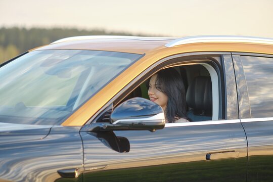 A woman with dark hair is seated in the driver's seat of a modern car, smiling as she enjoys a sunny day. The lush greenery and clear sky enhance the relaxed atmosphere of her drive