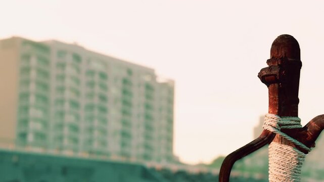 A rusty sword is tied with rope and stands upright. The background shows buildings under a clear sky during sunset. The scene captures the contrast of ancient and modern.