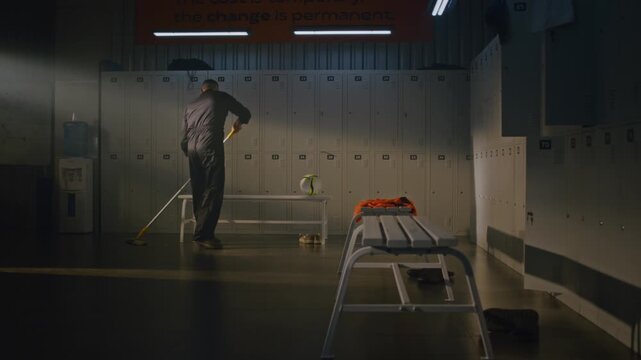 Professional Male Cleaner in Black Overalls is Methodically Mopping Floor of Empty Locker Room. Man Stops Working, Picks Up Left Behind Soccer Ball, Starts Skillfully Juggling it With Feet and Knees.
