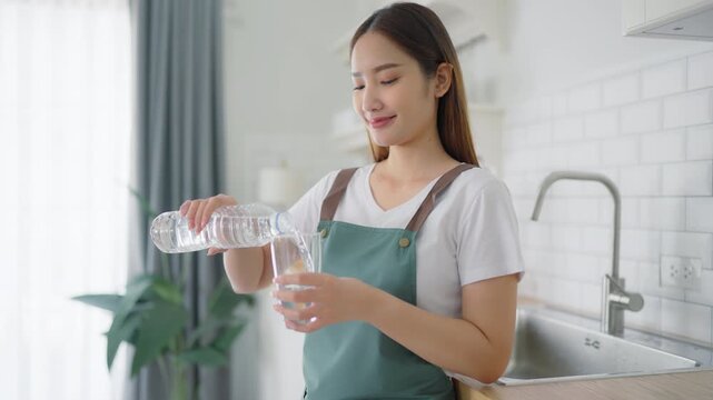 Happy young Asian woman drinking water while standing in the kitchen room. Healthy food and wellness lifestyle