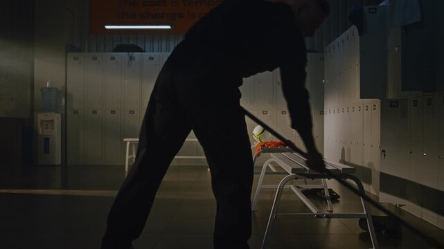 Focused Male Custodian Performing Structured Cleaning Routine. Man Carefully Mops Floor Around Left Behind Sports Gear and Systematically Inspects Rows of Lockers. Professional Doing Job Thoroughly.