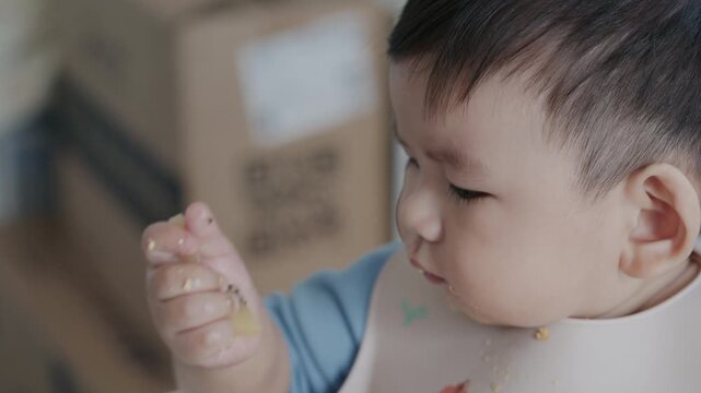 4K close up Cute Asian baby boy sitting in high chair and eating food with his hand at home.	