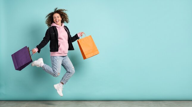 Excited woman jumping with shopping bags against a teal background, celebrating retail therapy and consumerism