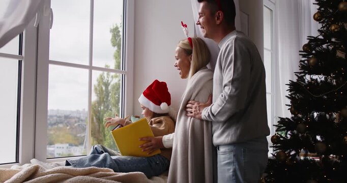 Christmas family with daughter looking at window at home. Parents cuddle near a tree the child in a Santa hat reading a book on the sill, gazing at winter streets. Winter holiday leisure together.
