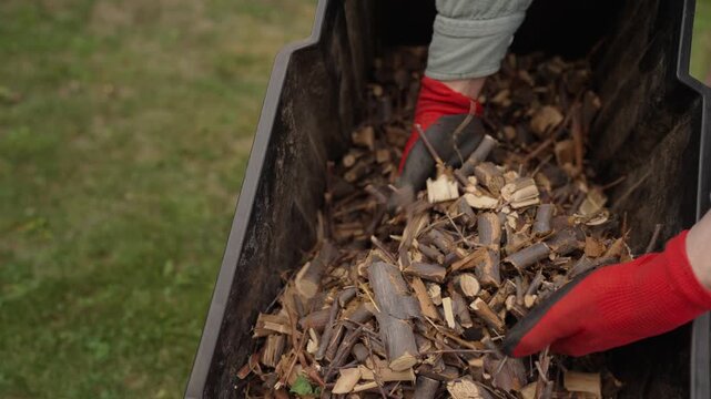 Person wearing red gloves handles fresh wood chips in a black container outdoors. Garden mulching and composting work in progress.