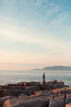 A person in traditional Chinese clothing looking back while standing on the rocky coast at sunset, profile view.