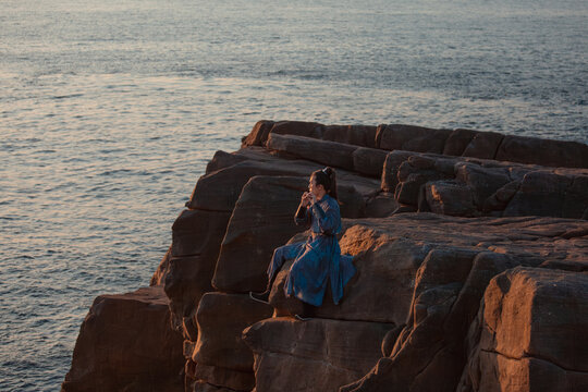 A person in traditional Chinese clothing plays a wind instrument while sitting on a rocky cliff by the sea at sunset, side view.