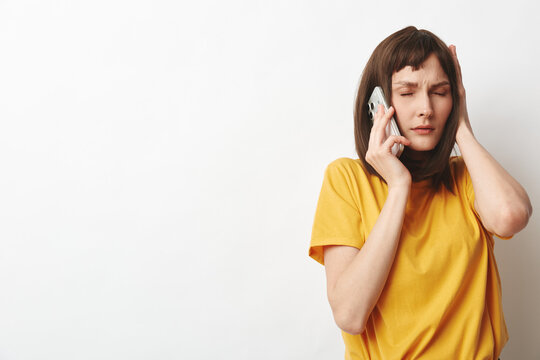 A stressed young woman in a yellow t shirt speaks on a cellphone, eyes closed and hand pressed to her temple, standing against a plain white wall in a candid moment.