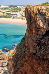 Dramatic layered cliffs overlook crystal-clear turquoise waters and a golden sandy beach along Portugal's Algarve coastline. A small coastal town with a lighthouse rises in the distant background © Aimur