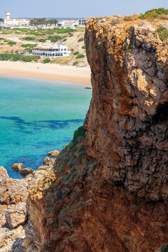 Dramatic layered cliffs overlook crystal-clear turquoise waters and a golden sandy beach along Portugal's Algarve coastline. A small coastal town with a lighthouse rises in the distant background