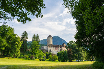 An ancient stone castle and round watchtower rise above a lush green valley surrounded by mountains in Austria. Manicured lawns and towering trees frame this picturesque historic landmark scene © Aimur