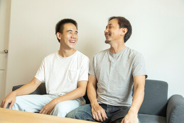 Obraz premium Happy Asian father and son sitting on sofa at home. Cheerful father with teenage son sitting on couch in the living room of apartment. Portrait of father and son spending leisure time at home