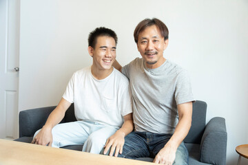 Obraz premium Happy Asian father and son sitting on sofa at home. Cheerful father with teenage son sitting on couch in the living room of apartment. Portrait of father and son spending leisure time at home