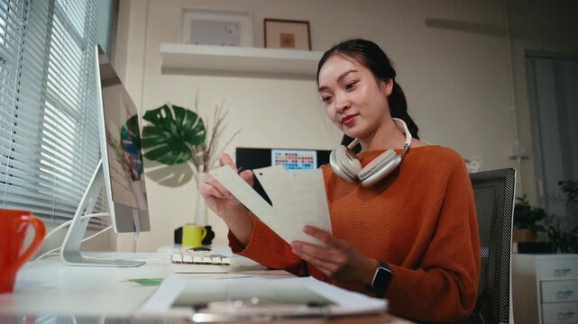 Focused Asian creative worker sitting at her desk, comparing color swatches with her computer screen while designing a new project at night
