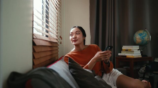 Young happy asian woman relaxing on a comfortable sofa, smiling while texting on her mobile phone and looking thoughtfully out the window
