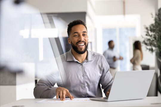 Laughing businessman sits at desk with laptop and printed charts, engaged in productive workflow, enjoy completion of successful project, received positive outcome. Career, professional opportunities