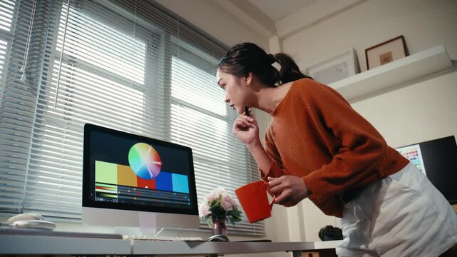 Young Asian female professional video editor working with a color wheel on a modern computer while drinking coffee in her home office