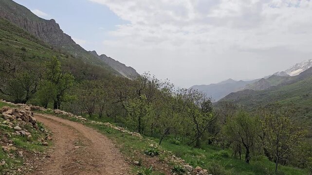 Spring mountain valley with lush green hills, snow-capped peaks, traditional stone wall, orchard trees, dirt road, and dramatic cloudy sky.
📍 Hawraman Takht, Kurdistan, Iran 