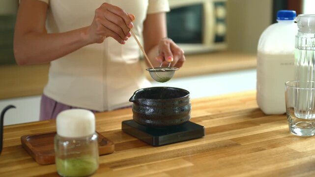 Closeup Woman uses bamboo scoop to sift vibrant green matcha powder through a fine mesh strainer into a dark ceramic