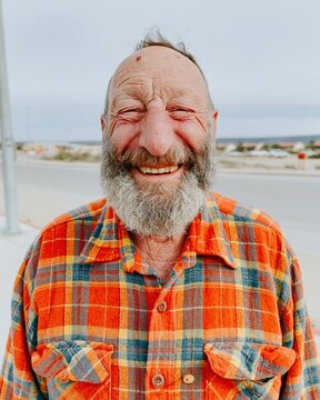 Smiling senior man with a grey beard wearing an orange plaid flannel shirt outdoors, showing a genuine warm expression for elderly lifestyle and retirement concepts