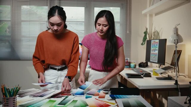 Two asian women graphic designers brainstorming and collaborating, choosing colors from swatches on their office desk for a new project