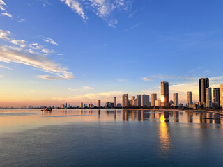 Fototapeta premium The sky glows a soft orange over the calm waters. The city skyline is reflected in the still, mirror-like surface. Clouds drift slowly across the deep blue expanse. Modern towers stand tall in the fad
