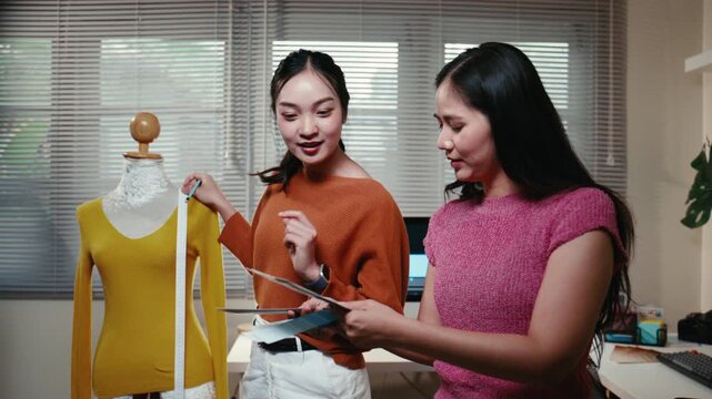 Two professional asian women working together in a design studio. One stylist measures a garment on a mannequin while her colleague chooses colors