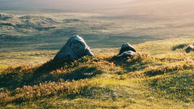 Two large rocks sit on green grass as the sun sets in the distance. The landscape has distant hills and soft shadows, showing the fading light of day.