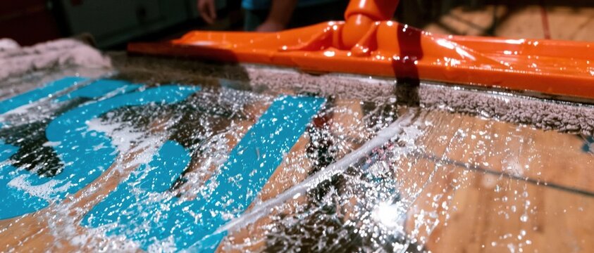 Orange floor squeegee spreading cleaning solution over a glossy gym floor, showing wet textures and reflections for facility maintenance and school renovation