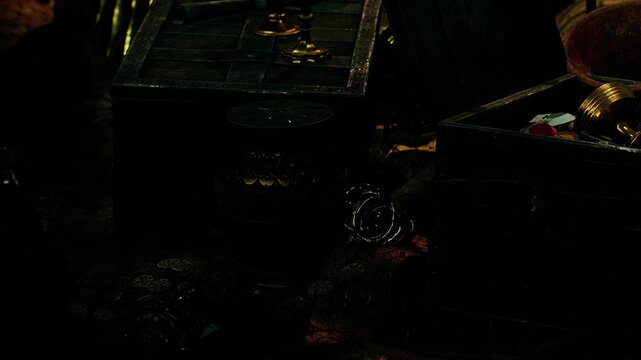 Treasure chests are open in a dark room. Gold coins and colorful jewelry pieces are scattered around. Some objects shine in the low light while others remain hidden in shadow.